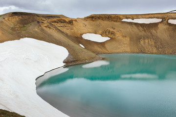 Viti crater lake with turquoise water surrounded by brown mountain slope covered by snow at Krafla volcano geothermal area in Iceland on a cloudy summer day