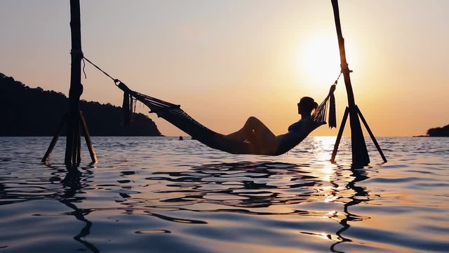 Woman relaxing in hammock at vacation holidays tropical beach destination with sunset light, silhouette of girl above sea water in paradise bay resort with scenic landscape