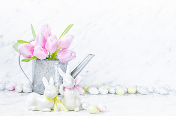 Easter table with porcelain bunnies, eggs and metal watering can with pink tulips on marble background.