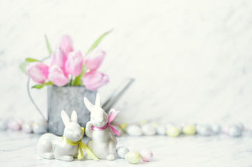 Easter table with porcelain bunnies, eggs and metal watering can with pink tulips on marble background.