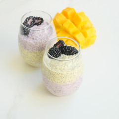 Chia seeds pudding with mango puree in beautiful glasses with blueberries and cut fresh ripe tropical fruit on light background - raw vegetarian sweet organic dessert.