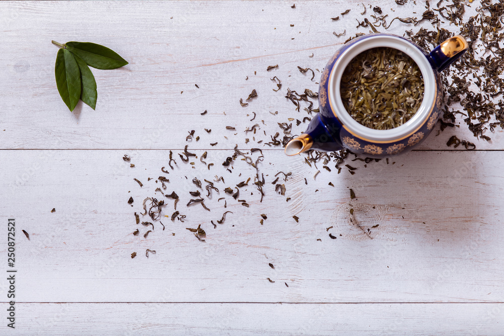 Wall mural Teapot and dry tea leaves on white wooden background, green tea leaf on table, black herbal homemade hot beverage in porcelain pot, english tea ceremony background, top view from above, copy space