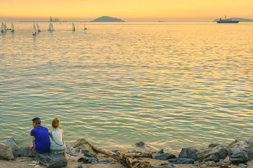 Back view of young couple sitting together on stone in front of ocean enjoying sunset during summer vacation.