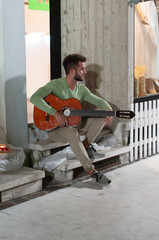 Young boy sitting on steps, with a beard, green shirt and beige trousers with sneakers while he plays the guitar. Concept of youthful carefree.