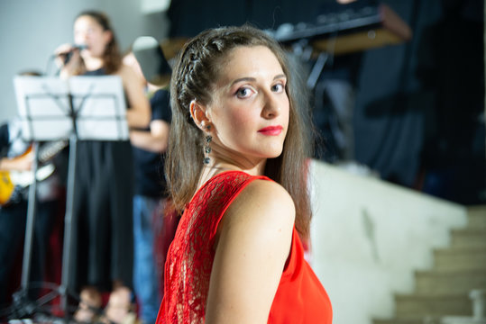 Young model sitting behind a musical complex. Girl in elegant dress with wide trousers and long lace insert on the back. Perfect makeup with red lips, hairstyle with braided hair.