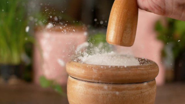 MACRO, DOF, SLOW MOTION: Unrecognizable woman crushes a chunk of rock salt sitting inside a mortar with a wooden pestle. Female chef strikes the clump of sea salt while gripping a round wooden stick.