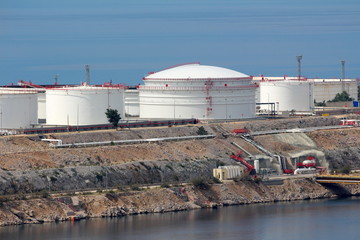 Freshly painted new large industrial white metal oil refinery tanks on top of rocky sea shore connected with multiple metal pipes to pier for unloading purpose on warm sunny day