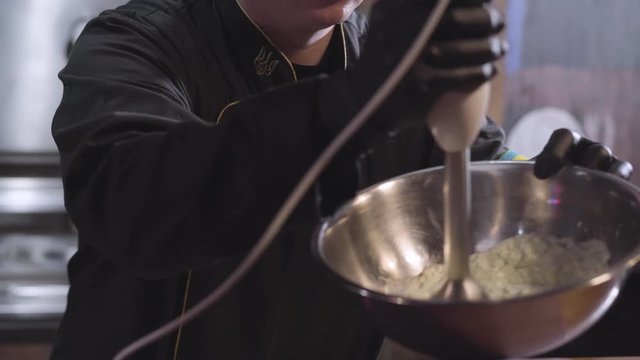 Chef in uniform and black rubber cook gloves whipping sause with blender in big aluminium bowl close up. Preparing food in modern restaurant