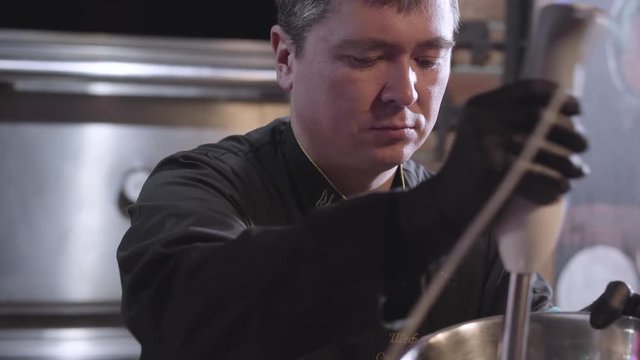 Chef in uniform and black rubber cook gloves whipping sause with blender in big aluminium bowl close up. Preparing food in modern restaurant