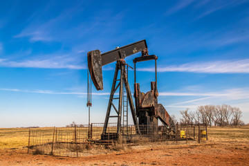 Large old rusty oil well pump jack surrounded by cattle panel fence out in field with very blue sky...