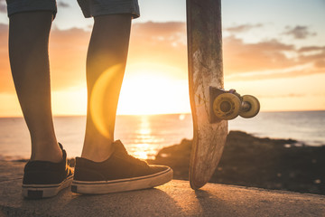 Teenager looking at sunset on the horizon holding a skateboard in hand. Teen wearing plaid shirt, shorts and black sneakers the sun sets, golden reflections on the shore of the ocean. Back close up © Fabio Principe