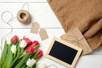 Bouquet of tulips flowers, blank picture frame, tags, twine, burlap on white wooden table. Vintage greeting card for womans day, mothers day, birthday, easter. Rustic background, flat lay, above view.