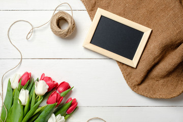  Bouquet of tulips flowers, blank picture frame, twine, burlap on white wooden table. Vintage greeting card for womans day, mothers day, birthday, easter. Rustic background, flat lay, above view.