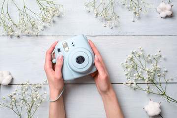 Female hands holding modern polaroid camera on a blue wooden background with flowers. Top view,...