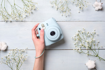Female hand holding modern polaroid camera on a blue wooden background with flowers. Top view,...