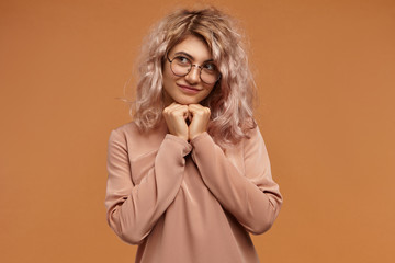 Adorable timid young woman wearing trendy round eyeglasses, holding clasped hands under her chin and looking away, having shy mysterious smile. Cuteness, youth, beauty, style and fashion concept