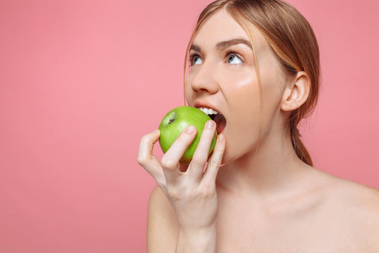 Portrait Of A Happy Beautiful Woman Holding An Apple Trying To Bite Him, With Healthy Teeth On A Pink Background