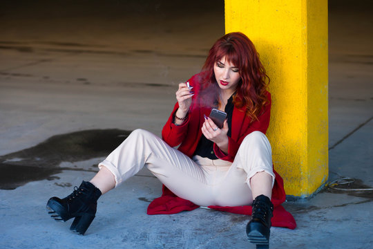 Smoking girl sitting with a phone in the parking. Young woman in red coat with a cigarette and smartphone in the underground parking