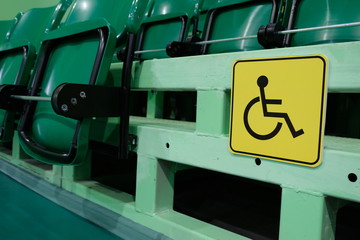 Yellow sign is a place for disabled people in the gym among the seats for spectators. Auditorium with rows of raised green chairs. Social sphere.