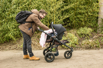 Bearded smiling father with a backpack and in yellow boots walking with a pram in a spring park shows care and love by covering a small child with a blanket.