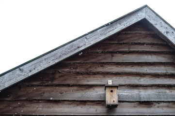 Birdhouse is located under the roof of a wooden house, against the background of a cloudy sky and snowflakes.