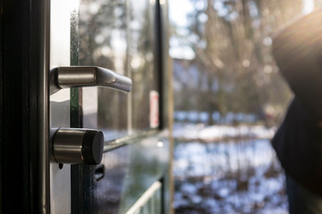 Metal entrance door with glass and a chrome scratched handle is open in the bright rays of the spring sun, against a blurred background of a snow-covered yard.