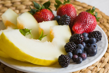 Sliced fruits and berries on a beautiful white plate. Melon, Strawberry, Blueberry, Blackberry and Mint Leaves. Dietary fruit salad for breakfast, weight loss concept. Healthy snack.