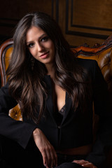 Lovely tanned girl with long shiny hair wearing black jacket posing on a vintage sofa
