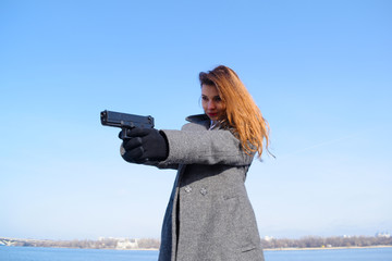 Young serious girl in a gray coat, shooting with a gun near the river in the city.