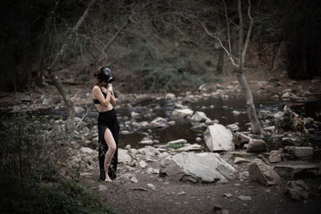 Obraz premium Side view of anonymous barefoot woman in black clothes and gas mask standing in amazing spooky forest