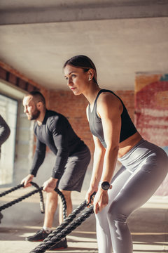 Caucasian fit couple exercising with battle ropes at gym. Woman and man dressed in sports outfit training together doing battling rope workout, with powder and dust particles flying in the sun rays