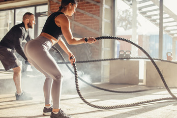 Active tense cross fit workout of a young caucasian couple in excellent physical shape, exercising with ropes in gym with panoramic windows and sun rays