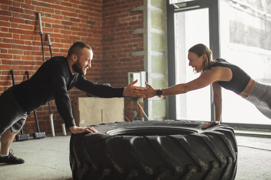 Caucasian crossfit male coach shows crossfit plyometric push-up technique, performed on truck tire to his female trainee , encouraging her with hand.