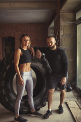 Young trained woman in sports outfit posing with her personal crossfit trainer, posing against truck tire in functional training studio