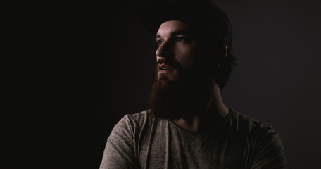 Fototapeta premium Handsome bearded man in tshirt and hat with crossed arms looking away on darkling background
