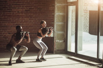 Young mixed-race fit couple doing squats exercise, while holding in hands heavy sandbag during...