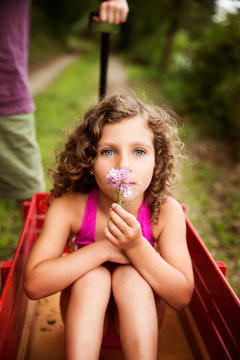 Girl (8-9) with curly hair sitting in red basket and holding pink flower person in background 