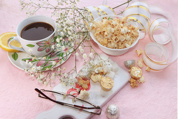 A cup of coffee with lemon and flowers in a festive ribbon, a plate with sweets on a pink background
