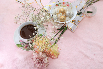 A cup of coffee and flowers in a festive ribbon, a plate with sweets on a pink background
