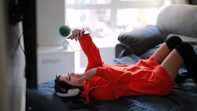 Young sexy girl talks by smartphone, online video chatting, communicate with friend lying at home bed
