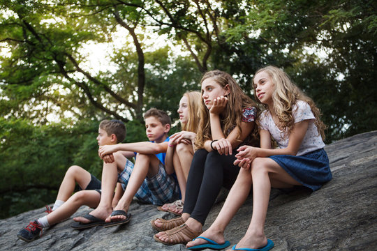 Children (8-9, 12-13, 14-15) sitting on rock in park 