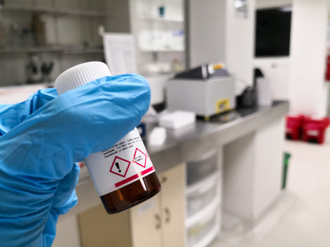 Hand with a safety glove holding a chemical reagent in an amber bottle. Background of a scientific research laboratory.