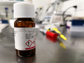 Chemical in a small amber bottle. Scientific laboratory with equipment, pipettes and markers in the background.