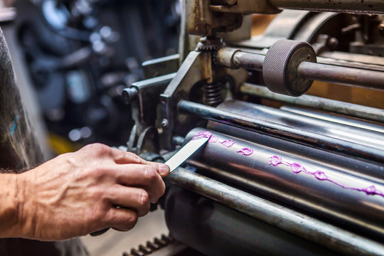 Hand with utility knife applying ink to printing press 