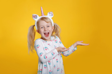Cute adorable smiling Caucasian blonde girl in white dress wearing unicorn headband horn and ears in studio on yellow background. Funny kid child expressing emotion and showing pointing