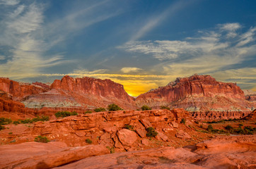 Sunset during golden hour in Southern Utah, sun warming red sandstone, cliffs, mountains, and mesa. Sun is peaking out behind the rocky peaks