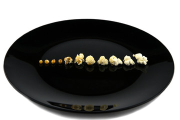 popcorn cooked in the microwave at home: the Different stages of the cooking of corn grains laid out on a black plate in a row, side view  isolated on white