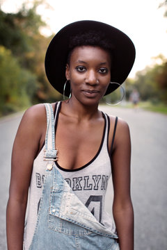 Portrait of young female wearing earrings, hat, tank top and denim jeans with suspenders 