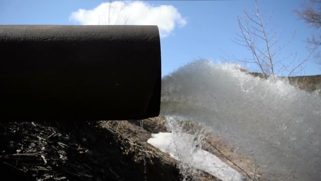 A black storm drain pipe with storm water runoff flowing out of it into a spillway leaching into the local water table