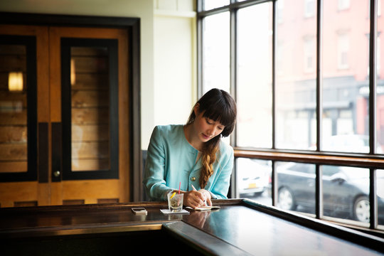 Young woman sitting at bar counter with drink and writing in notepad 